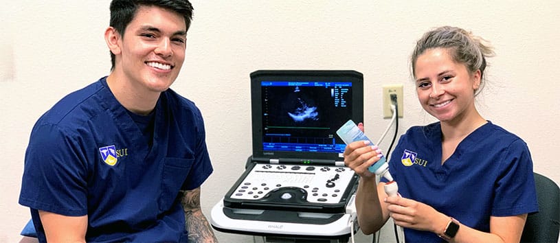 Two SUI Sonography students wearing blue scrubs and smiling in front of a sonography screen.
